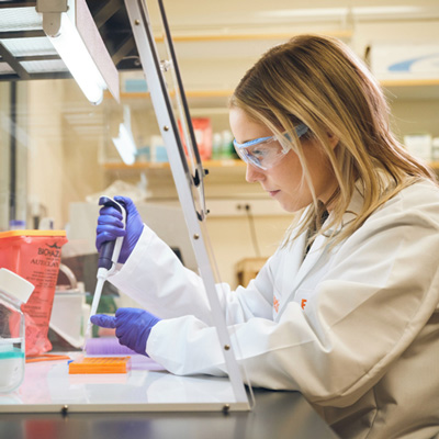 Zoetis scientist in a lab coat and blue gloves uses a pipette, with sample tubes and containers on the work surface. - Zoetis