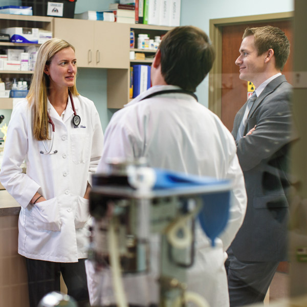 A veterinarian in a white coat with a stethoscope speaks to two people, with medical equipment and shelves in the background - Zoetis